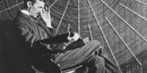 Nikola Tesla, with Rudjer Boscovich's book "Theoria Philosophiae Naturalis", in front of the spiral coil of his high-voltage Tesla coil transformer at his East Houston St., New York, laboratory.